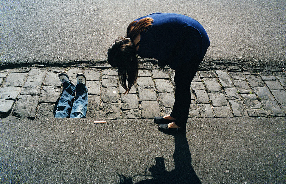 Eine Frau beugt sich und zwei Beine schauen unter Straße hervor. Melbourne, Straßenfotografie von Jesse Marlow.