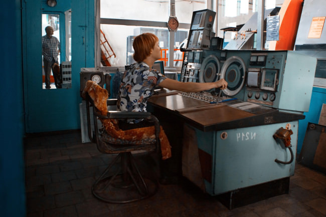 Eine Frau mit roten Haaren und Kurzhaarschnitt sitzt an alten Gerätschaften einer Industrieanlage.