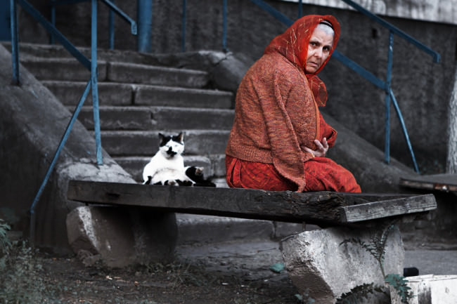 Eine alte Frau sitzt mit einer Katze auf einer Steinbank und dreht sich zum Betrachter.
