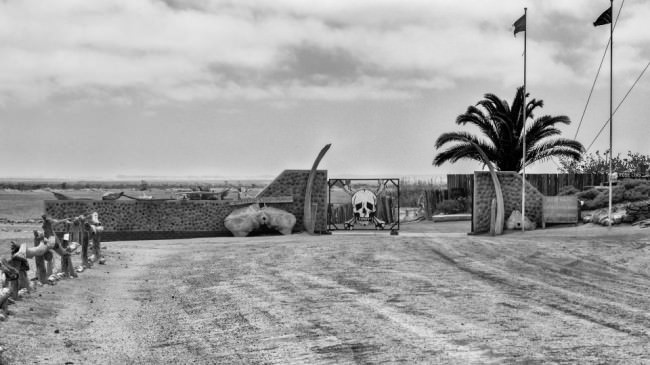 Einfahrt zu einem Hof in der Wüste, am Tor das große Bild eines Totenschädels.