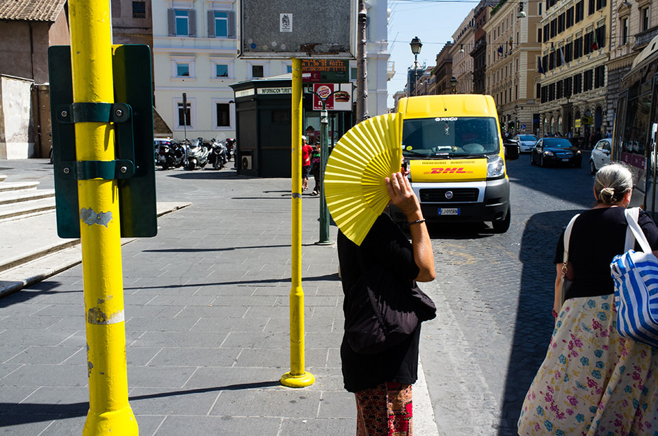 Eine Frau mit gelbem Fächer steht an der Straße und hält sich den Fächer vor das Gesicht. 