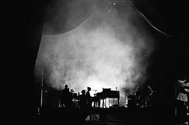 Blick auf die Bühne im Gegenlicht mit viel Rauch und einem Klavier sowie Menschen.