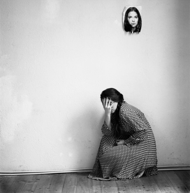 Eine Frau hockt auf dem Boden und versteckt ihr Gesicht. An der Wand hängt ein Bild von ihr.