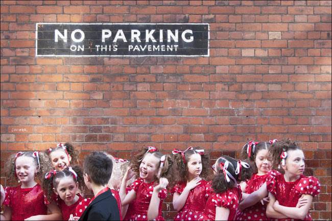 Viele Mädchen in roten Kleidern und ein Junge im Anzug vor einer Backsteinmauer mit der Aufschrift „No parking on this pavement“.