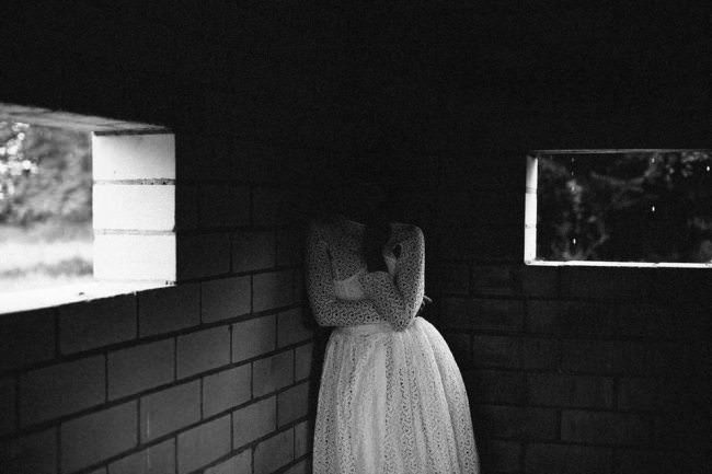 Eine Frau im weißen Kleid in einer Ecke eines Gebäudes. Ihr Gesicht verschwindet in der Dunkelheit.