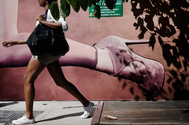Eine Frau in Turnschuhen passiert eine Wand mit großformatiger Werbung für hochhackige Schuhe.