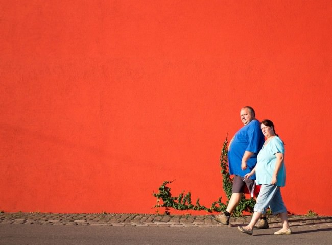 Ein Mann und eine Frau gehen vor einer knallroten Wand mit etwas Efeu vorüber.
