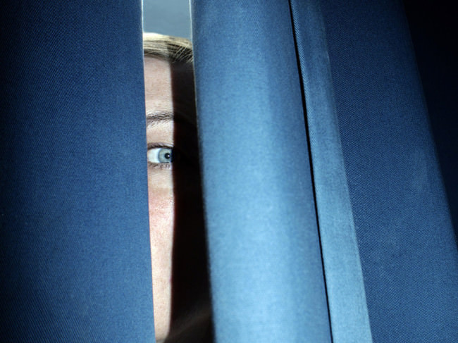 Im Spalt zwischen blauen Vorhängen ist das Auge einer blonden Frau zu sehen.
