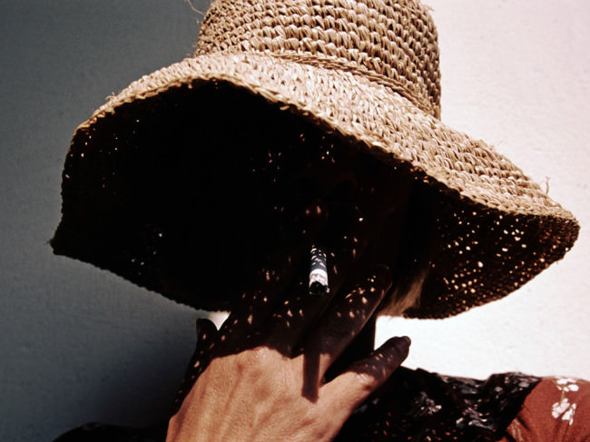 Eine Person hält sich eine Zigarette an den Mund, ihr Gesicht liegt im Schatten eines Strohhuts.