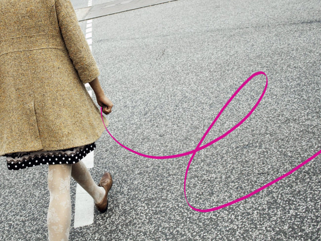 Eine Frau mit gepunktetem Rock, weißer Spitzen-Feinstrumpfhose und Mantel überquert eine Straße, digital wurde ihr ein gewundenes, pinkes Band an die Hand gegeben.