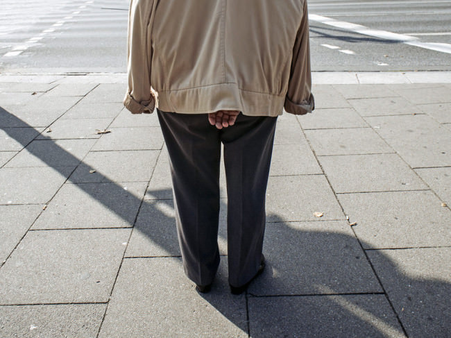 Rückansicht eines Mannes, der auf dem Bürgersteig steht und die Hände unter der Jacke hinter seinem Rücken verschränkt hält.