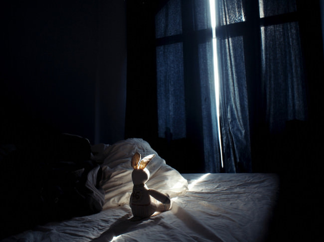 Auf einem Bett sitzt ein Plüschtierhase, der von einem Lichtstrahl erhellt wird, der durch einen Spalt zwischen Vorhängen in das sonst dunkle Zimmer fällt.