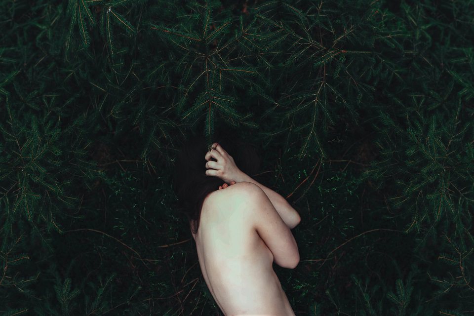 Eine Frau liegt in grünen Zweigen und umarmt sich selbst.