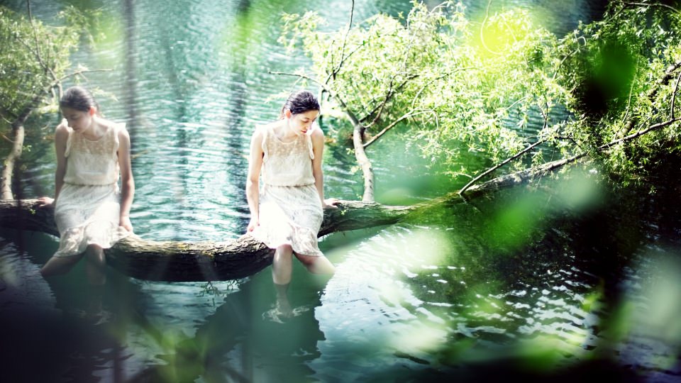 Ein Mädchen in weißem Kleid sitzt auf einem Baumstamm, der ins Wasser ragt.