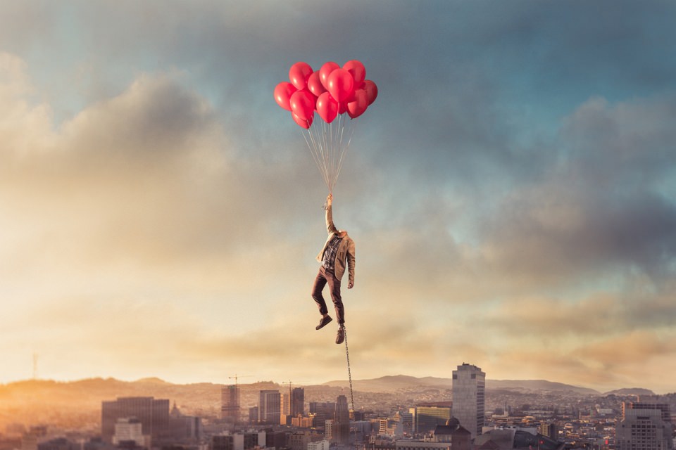 Ein kopfloser Mann schwebt an einem Strauß roter Luftballons über einer Stadt, an seinem Fuß führt eine Metallkette nach unten.