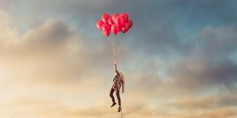 Ein kopfloser Mann schwebt an einem Strauß roter Luftballons über einer Stadt, an seinem Fuß führt eine Metallkette nach unten.