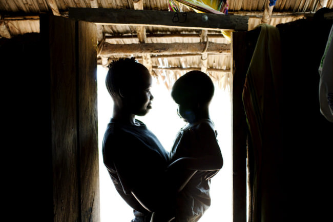 Eine Frau hält in einem Türrahmen ein Kind auf dem Arm. Sie stehen im grellen Gegenlicht, das von draußen in den dunklen Raum fällt.