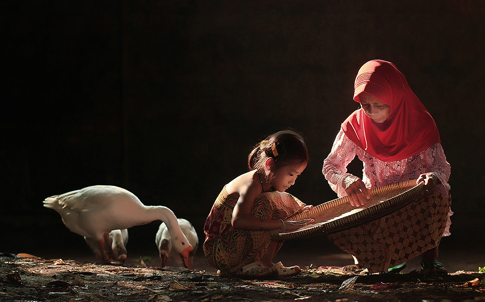 Eine Frau und ein Mädchen hocken mit einer großen Schale auf dem Boden neben zwei Gänsen.