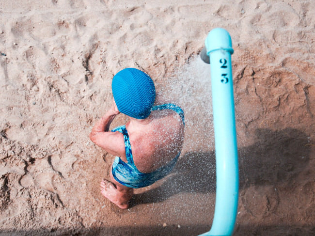 Eine Frau in blauem Badeanzug und mit blauer Badekappe unter einer Dusche am Strand.