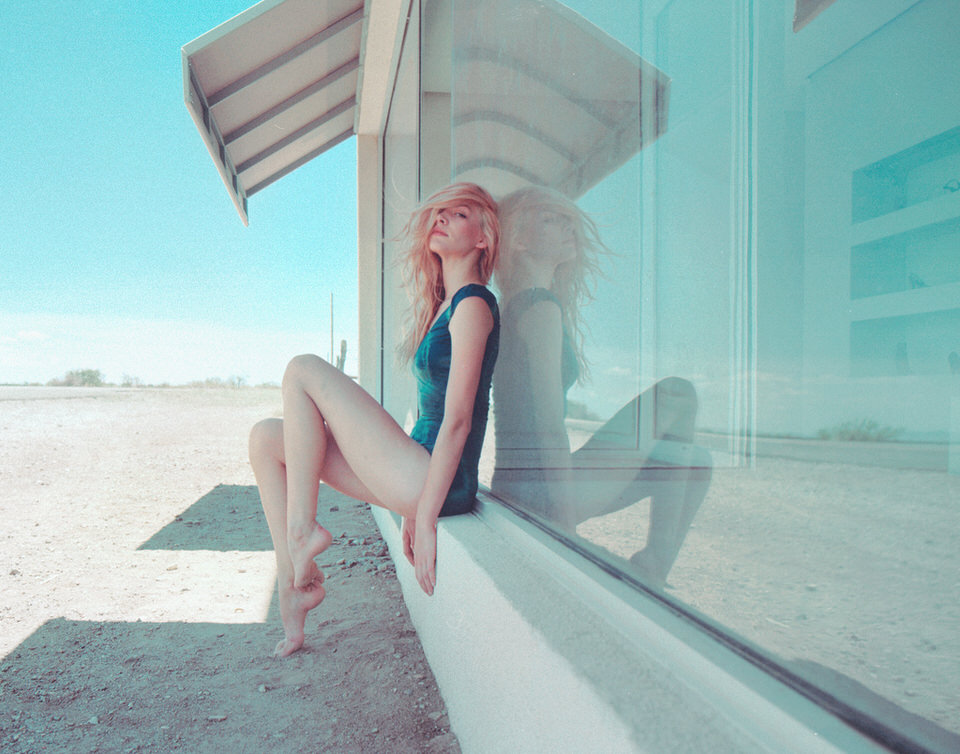 Blonde Frau in einem blauen Body vor einem großen Fenster im Nirgendwo.
