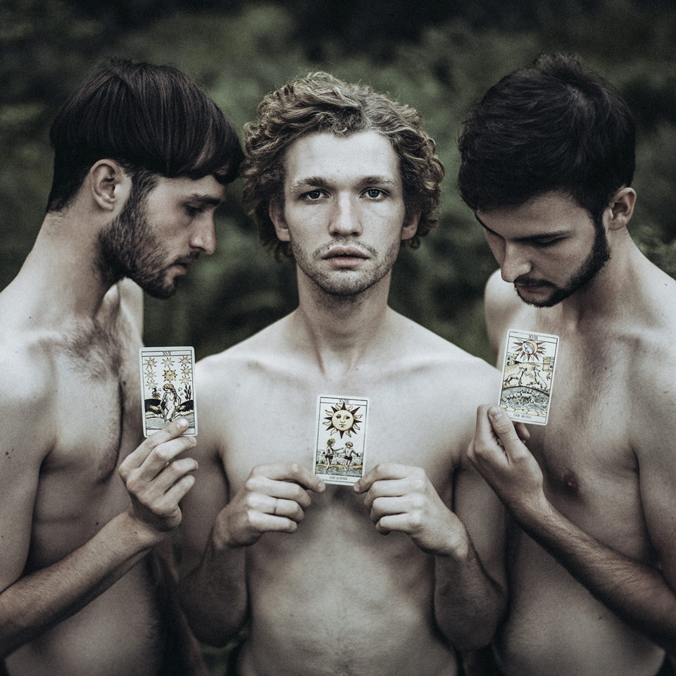 Drei Männer stehen nebeneinander und halten jeder eine Tarotkarte vor ihre Brust.