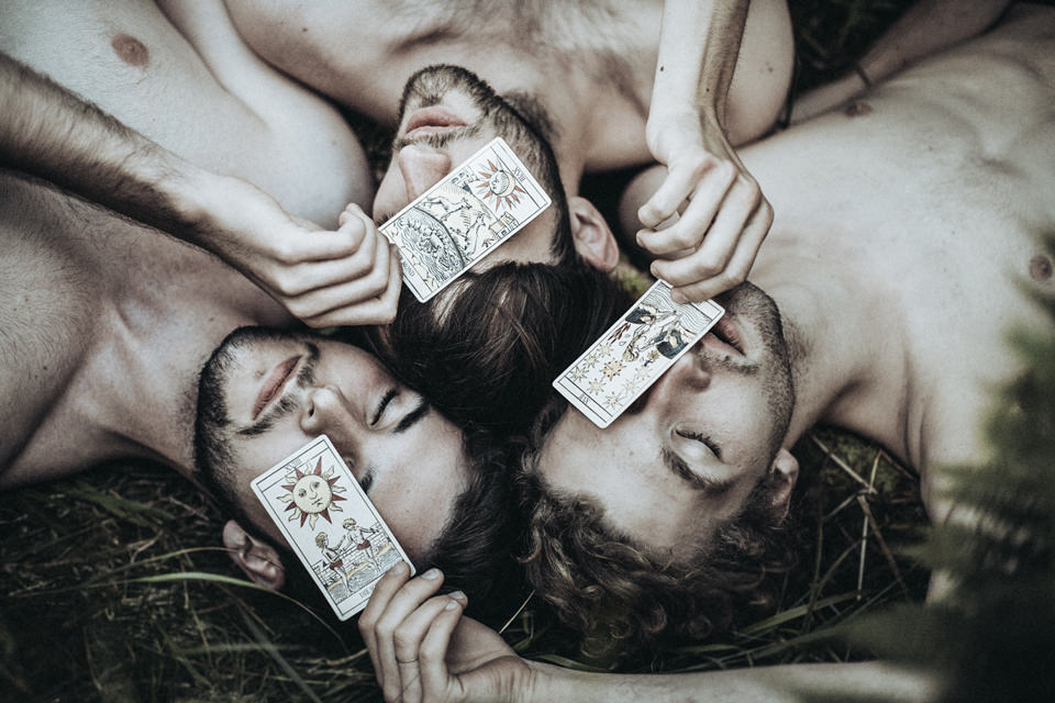 Drei Männer liegen auf dem Waldboden und decken gegenseitig ihre Augen mit Karten ab.