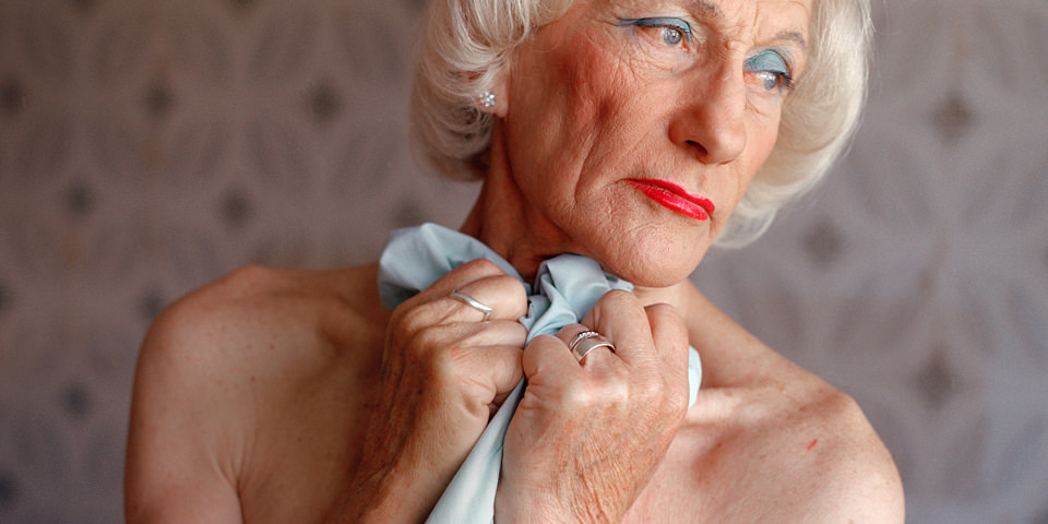 Eine ältere Frau mit weißen Haaren und rot geschminkten Lippen schaut versunken weg.