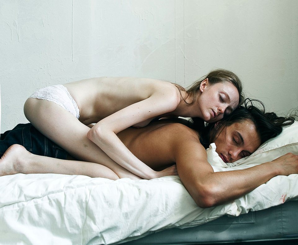 Eine Frau, nur mit Unterhose bekleidet, hockt auf dem Rücken eines Mannes auf einem Bett.