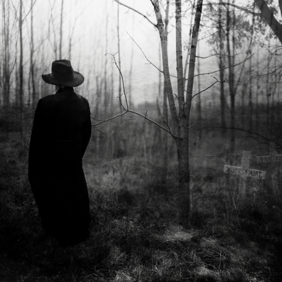 Ein Mann im schwarzen Mantel steht im Wald neben einem Kreuz.