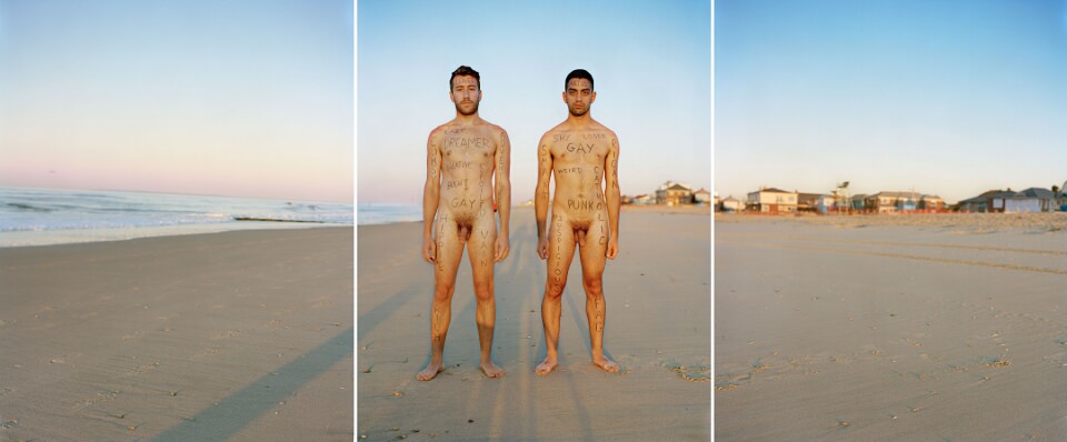 Zwei nackte und beschriftete Männer stehen am Strand.