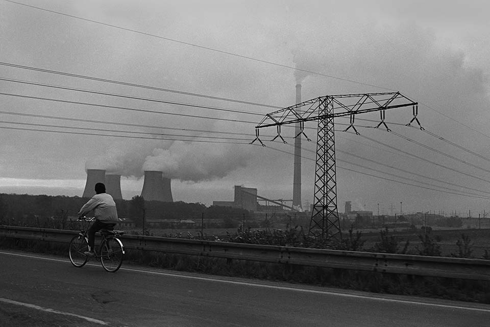 Ein Kohlekraftwerk stößt dicke Rauchschwaden in den bewölkten Himmel aus. Im Vordergrund ein Radfahrer.