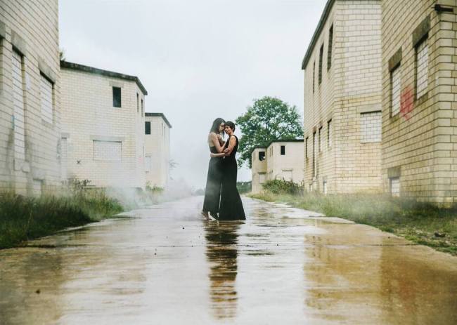 Zwei Frauen in schwarzen Kleidern umarmen sich im Regen zwischen zugemauerten Häusern.