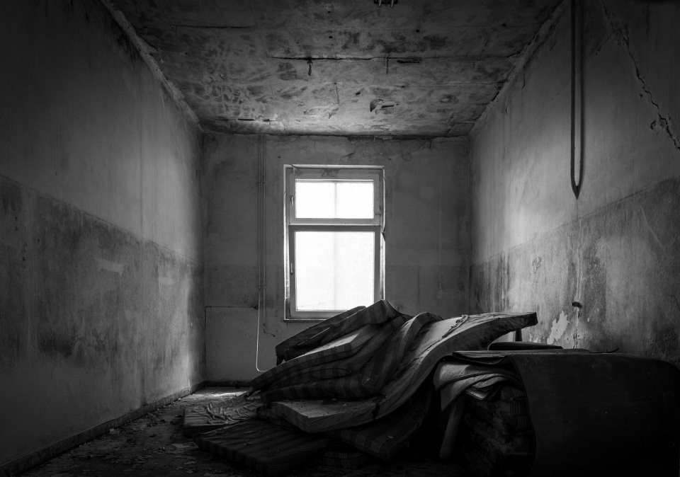 Stapel einiger Matratzen in einem verlassenen Raum; durch ein Fenster fällt Licht von hinten auf den Matratzenstapel.