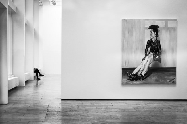 Ein Bild mit einer sitzenden Person im Museum, im Hintergrund sind die Beine eines sitzenden Besuchers zu sehen.