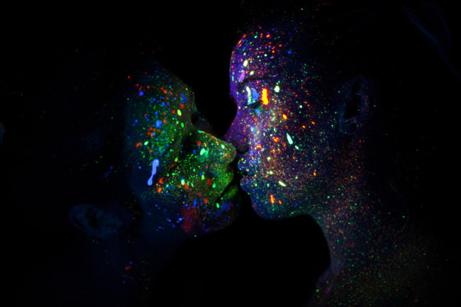 Zwei mit bunten, im UV-Licht strahlenden Punkten übersähte Personen, die sich küssen.