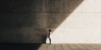 Eine Frau vor einer hohen Betonwand, halb im Licht, halb im Schatten stehend.