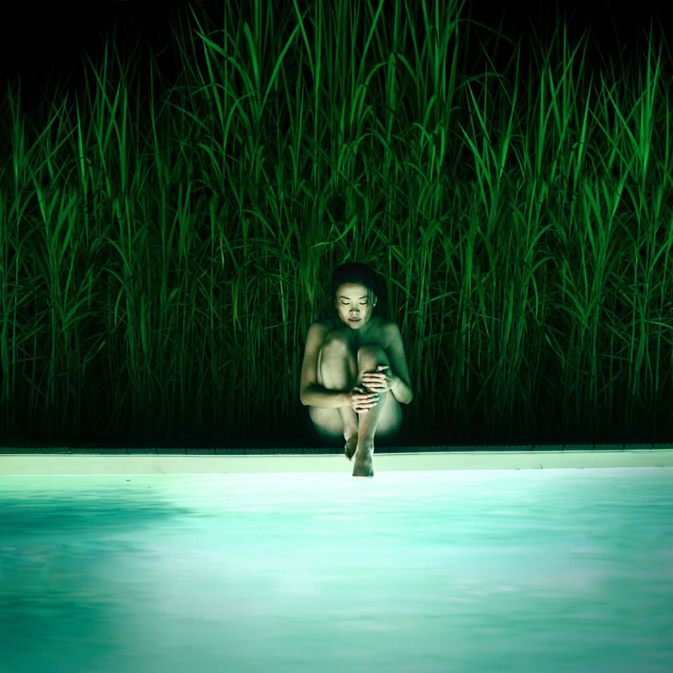 Nackte Frau sitzt mit angewinkelten Beinen am Rand eines beleuchteten Pools in der Dunkelheit, hinter ihr hohes grünes Gras.