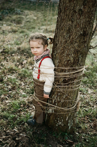 Ein Mädchen steht an einen Baum angebunden mit grimmiger Grimasse. 