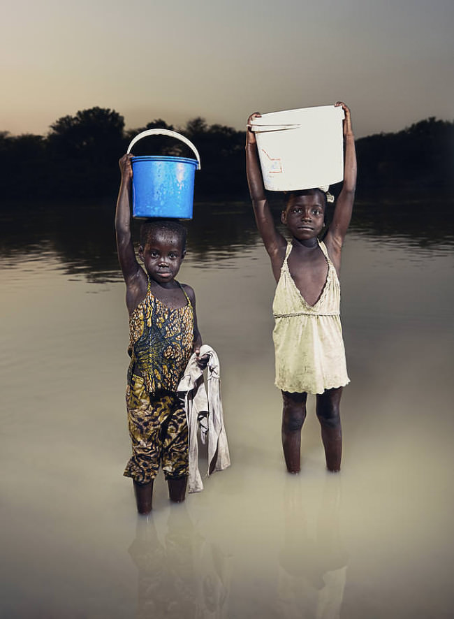 Zwei Kinder stehen im Wasser und tragen Eimer auf dem Kopf.