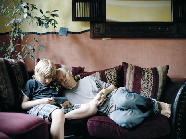 Ein Vater schläft auf der Couch an der Schulter eines Kindes, während das ein Videospiel spielt. 
