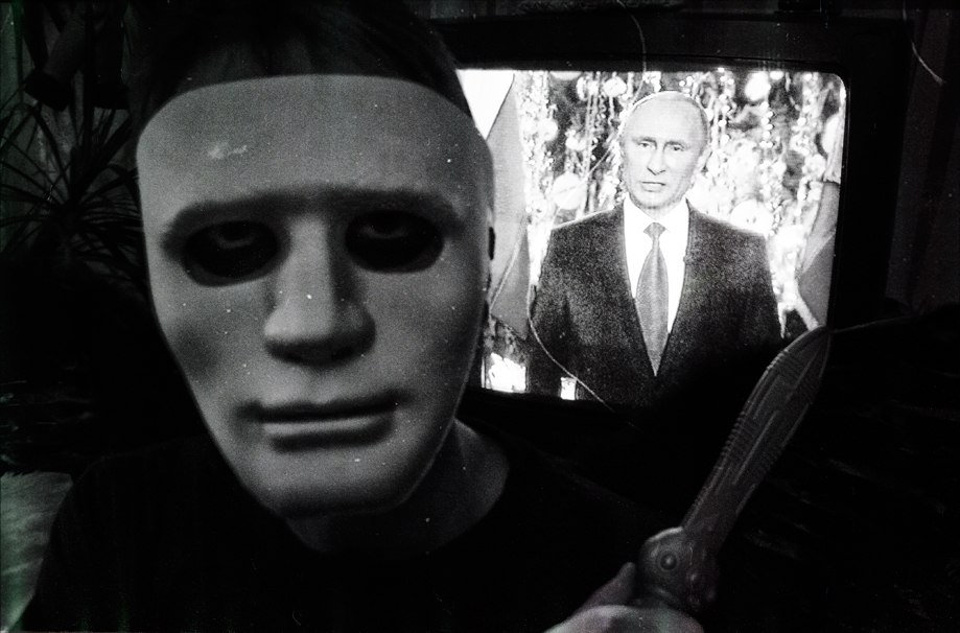 Ein Maskierter steht vor einem Fernseher, auf dem eine Rede von Putin läuft.