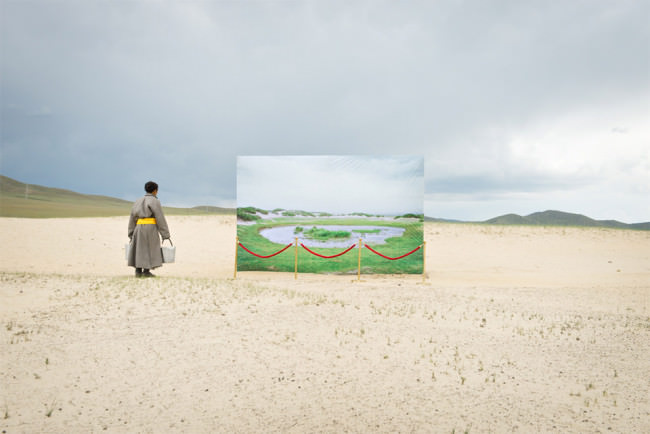 Ein Mensch mit zwei Eimern steht auf einer Sandebene vor einem Bild einer grünen Landschaft mit Wasserloch
