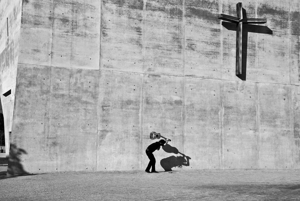 Ein Mann ist dabei, eine Gitarre auf den Boden vor einer Betonwand mit einem großen Kreuz zu schlagen.