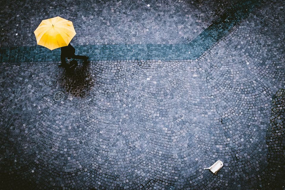 Mensch mit einem gelben Regenschirm auf Kopfsteinpflaster und etwas Weggeworfenes.
