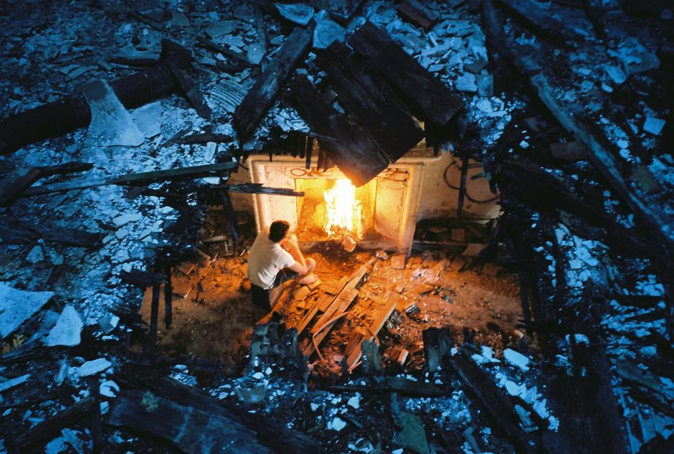 Ein Mann sitzt vor einem Kaminfeuer, betrachtet durch ein Loch in der Decke, bedeckt von Schnee und herumliegenden Holzbrettern, teilweise in den Raum darunter gefallen.