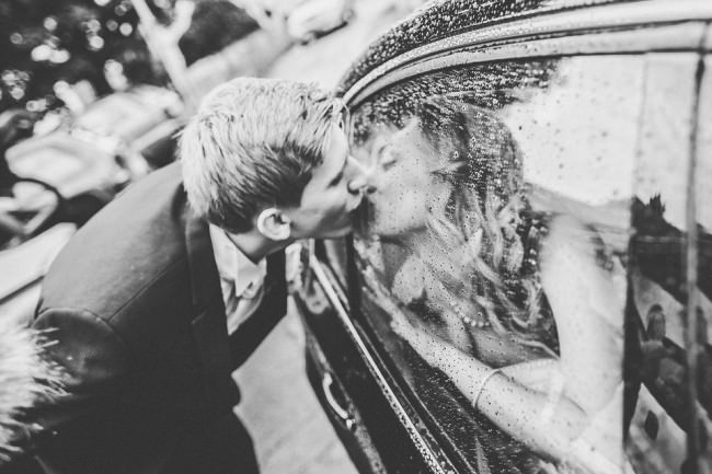 Ein Hochzeitspaar küsst sich durch eine Autoscheibe hindurch.