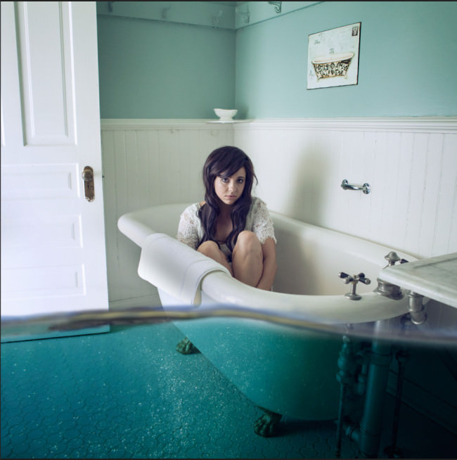 Eine Frau sitzt in einer leeren Badewanne, die von Wassermassen umgeben ist.