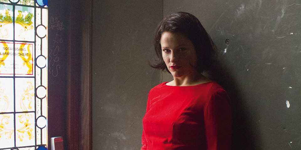 Portrait einer Frau mit rotem Oberteil vor dunkler Wand, links ein Fenster.