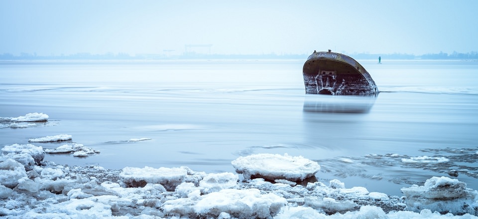 Schiffsrumpf ragt aus einem blauen Gewässer auf, am Ufer Schnee und Eis.