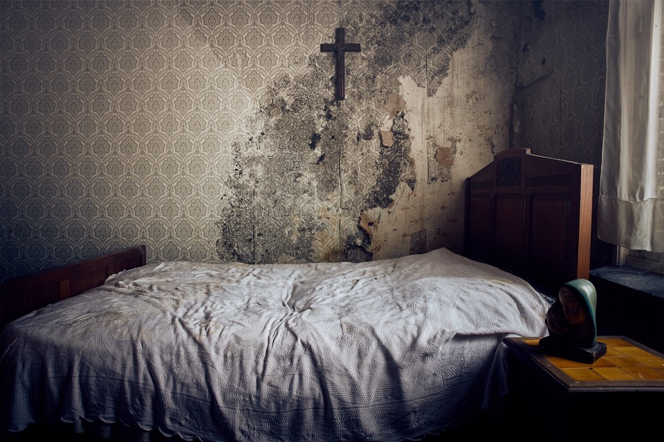 Schmutziges Bett vor einer verschimmelten Wand mit alter Tapete und einem Kreuz.